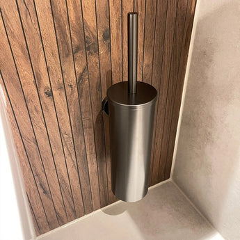 De Lauro Gunmetal WC-set van yessential.nl is een slanke metalen toiletborstelhouder met een verticale handgreep, gemonteerd op een wand met houten panelen naast lichtgekleurde tegels, perfect voor luxe badkamer accessoires collecties.