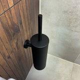 De Branco Black toiletborstelhouder van yessential.nl is gemonteerd op een wand met houten panelen naast beige tegels, wat een stijlvolle en praktische touch geeft aan het opbergen van je toiletborstel zwart.