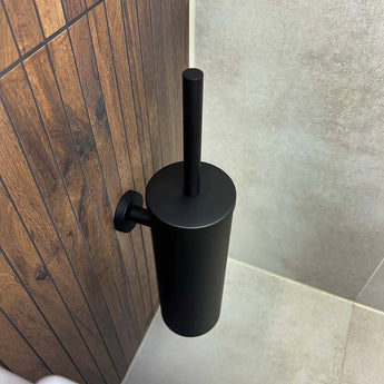 De Branco Black toiletborstelhouder van yessential.nl is gemonteerd op een wand met houten panelen naast beige tegels, wat een stijlvolle en praktische touch geeft aan het opbergen van je toiletborstel zwart.