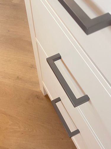 Close-up van een witte kast met drie lades, elk voorzien van Vasco Gunmetal handgrepen van yessential.nl. Geplaatst op een lichte houten vloer combineert dit meubel moderne stijl en functionaliteit voor elke kamer.