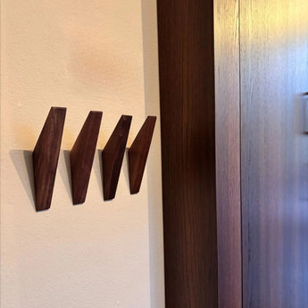 Drie moderne Fabio Walnut kapstokhaakjes van yessential.nl zijn gemonteerd op een beige muur naast donker hout, waardoor ze zachte schaduwen werpen en een minimalistische houten kapstok uitstraling creëren.