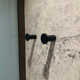 Vier zwarte Carreno haken van yessential.nl zijn gemonteerd op een muur met textuur en een steenpatroon. De haken werpen zachte schaduwen naar rechts en een glas of reflecterend oppervlak verschijnt gedeeltelijk aan de linkerkant van het beeld.