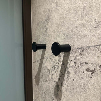 Vier zwarte Carreno haken van yessential.nl zijn gemonteerd op een muur met textuur en een steenpatroon. De haken werpen zachte schaduwen naar rechts en een glas of reflecterend oppervlak verschijnt gedeeltelijk aan de linkerkant van het beeld.